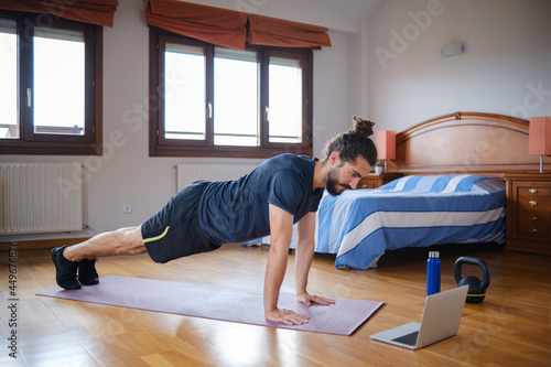 Bearded man doing plank while exercising at home with an online class on the laptop.