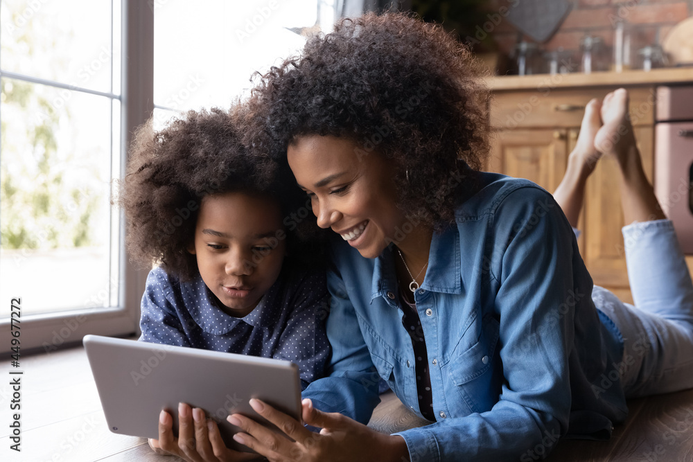Happy loving young African American mom and teen daughter relax on wooden kitchen floor have fun using tablet together. Smiling ethnic mom and biracial girl child speak on webcam video call on pad. Happy loving young African American mom and teen daughter relax on wooden kitchen floor have fun using tablet together. Smiling ethnic mom and biracial girl child speak on webcam video call on pad.