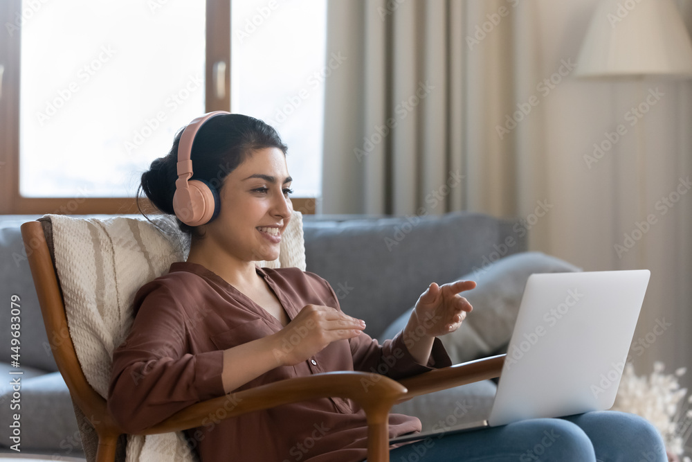 Smiling Indian woman in earphones sit in chair talk speak on video call on laptop. Happy mixed race ethnicity female in headphones have webcam digital virtual event on computer. Technology concept. Smiling Indian woman in earphones sit in chair talk speak on video call on laptop. Happy mixed race ethnicity female in headphones have webcam digital virtual event on computer. Technology concept.