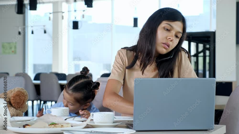 Mother feeds daughter in restaurant. Asian businesswoman mom gives pizza slice to toddler child sitting at table with tea and grey lapto and talking on black smartphone closeup Mother feeds daughter in restaurant. Asian businesswoman mom gives pizza slice to toddler child sitting at table with tea and grey lapto and talking on black smartphone closeup