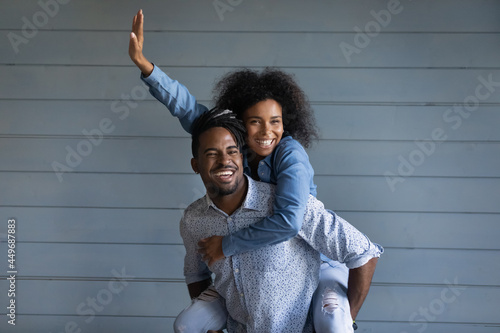 Overjoyed young African American couple renters have fun hug cuddle isolated on grey wall background. Smiling millennial ethnic woman piggyback happy biracial man show love. Rental, moving concept.