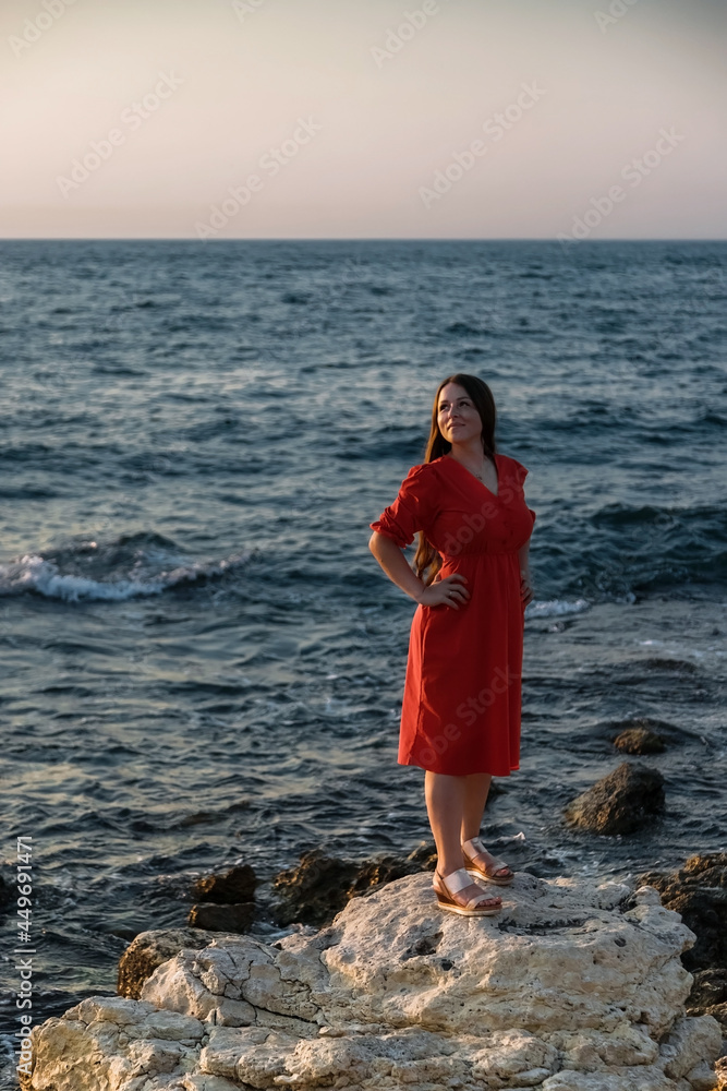 A girl in a red dress on the beach. The stone coast of the sea. Summer trip.