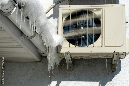 An outdoor air conditioner unit installed on the outer wall of a residential building close-up. Operation of the air conditioner in winter at low temperatures. Icicles and snow on the air conditioner