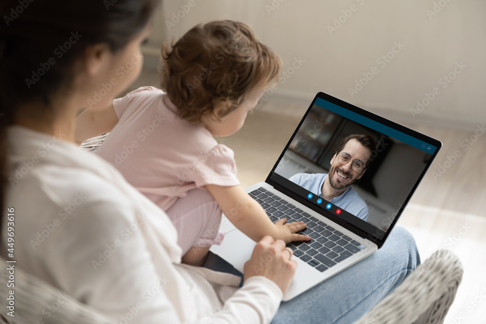 Over shoulder close up view of young Caucasian mom with toddler baby daughter speak talk on video call on laptop with smiling father. Mother with newborn kid have online communication on computer.