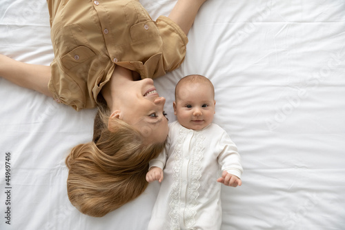 Top view portrait of happy young Caucasian mother and small baby infant kid lying in white bed at home relax together. Smiling mom and little newborn child rest in bedroom. Motherhood concept.