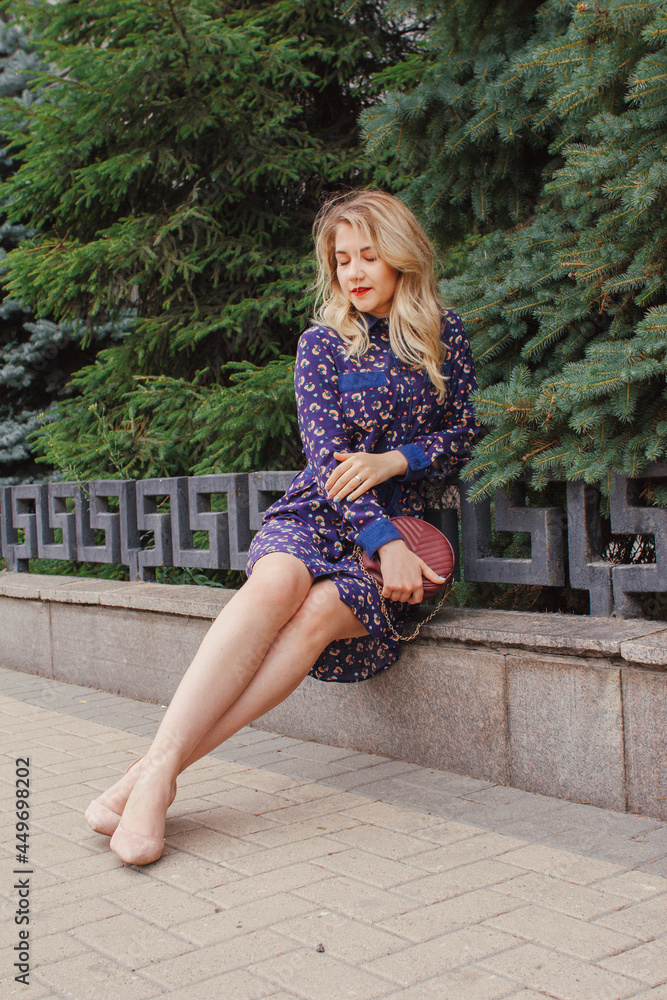 Naklejka premium Young beautiful woman in blue dress with a round bag walking on the spring street. Urban background. Stylish Tourist girl enjoying walking the city during weekend trip.