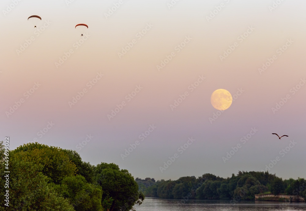 Paragliders with a motor and a seagull fly in the evening over the river against the background of the sky and the moon, landscape.