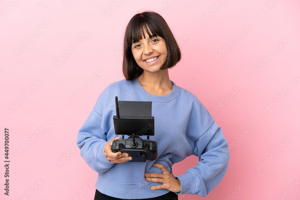 Young mixed race woman holding a drone remote control isolated on pink background posing with arms at hip and smiling Young mixed race woman holding a drone remote control isolated on pink background posing with arms at hip and smiling