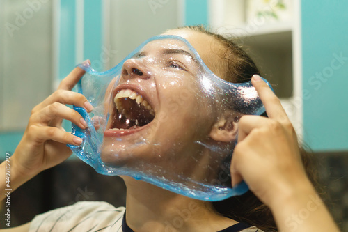 A cheerful, laughing girl looks through a transparent, stretched blue slime.Homemade slime on the face of a happy girl. A charming young woman is playing with a popular anti-stress toy.Selective focus
