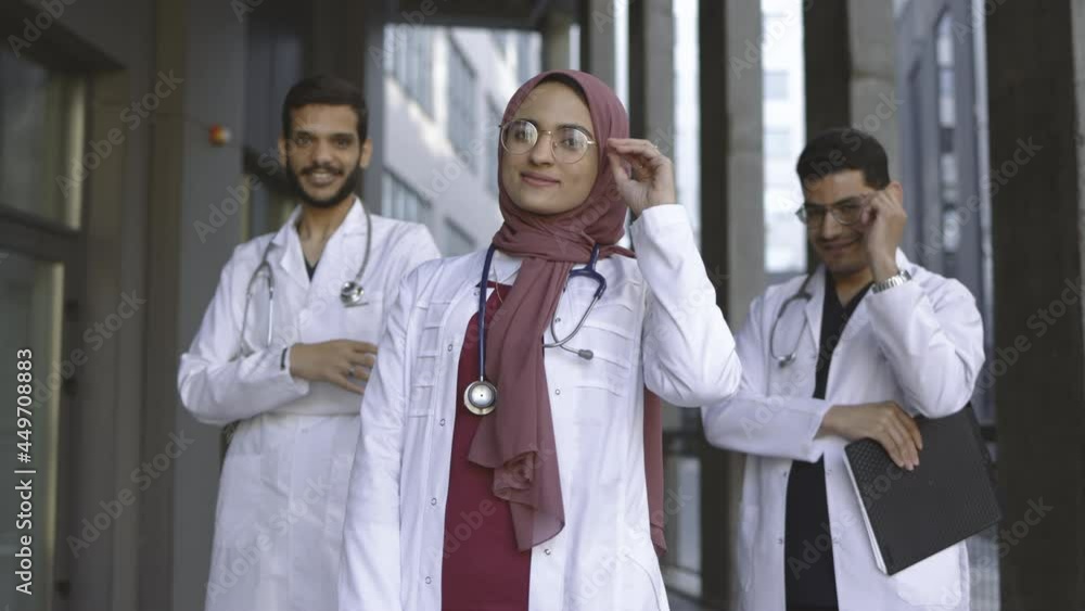 Young female Muslim medical student with her male groupmates behind, standing outdoors and smiling at camera, adjusting eyeglasses and showing ok gesture. Medical team outside hospital