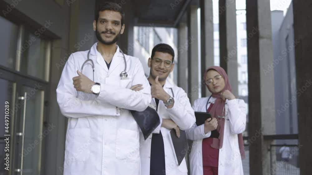 Portrait of three young Arabic Malay medical students team showing thumbs up while standing in a row outside modern clinic or university campus Portrait of three young Arabic Malay medical students team showing thumbs up while standing in a row outside modern clinic or university campus