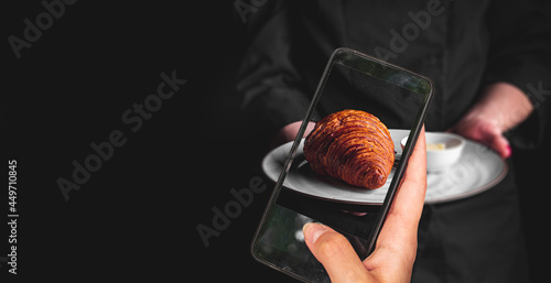 woman hand holding and showing smart phone takes a photo chef in a black suit holds in his hands plate with croissant on Dark grey black background. food photos