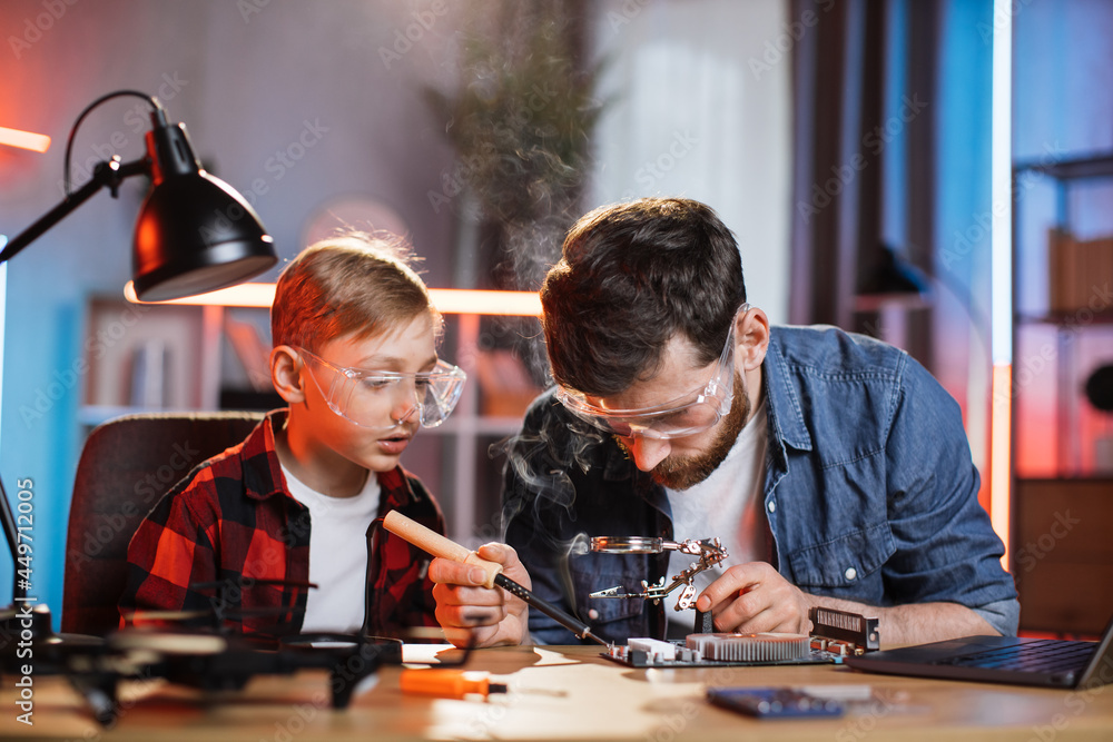 Fototapeta premium Caucasian father and his son in protective glasses using soldering iron for fixing display card from laptop. Two men sitting together at table and repairing modern technology at home.