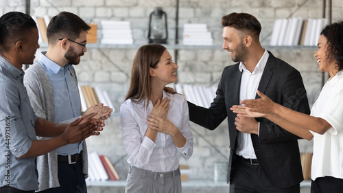 Smiling supportive diverse businesspeople congratulate excited young Caucasian female employee with job promotion or success. Happy multiethnic colleagues greeting woman worker with work achievement.