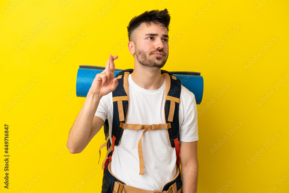 Young mountaineer caucasian man with a big backpack isolated on yellow background with fingers crossing and wishing the best Young mountaineer caucasian man with a big backpack isolated on yellow background with fingers crossing and wishing the best