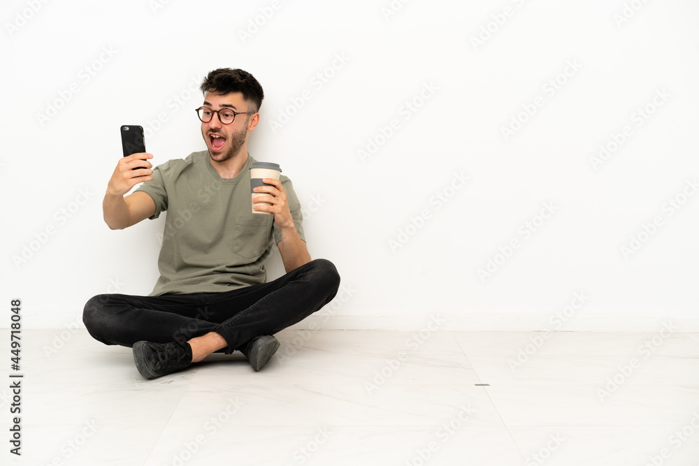 Young caucasian man sitting on the floor isolated on white background holding coffee to take away and a mobile Young caucasian man sitting on the floor isolated on white background holding coffee to take away and a mobile