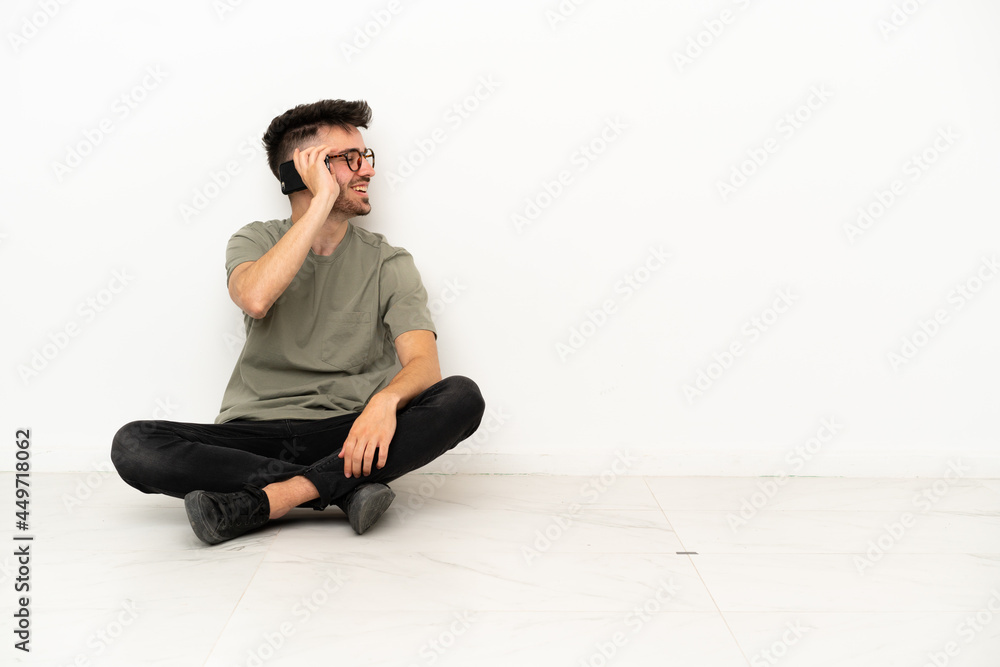 Young caucasian man sitting on the floor isolated on white background keeping a conversation with the mobile phone with someone Young caucasian man sitting on the floor isolated on white background keeping a conversation with the mobile phone with someone