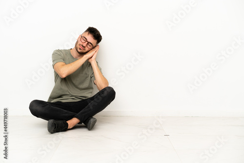 Young caucasian man sitting on the floor isolated on white background making sleep gesture in dorable expression