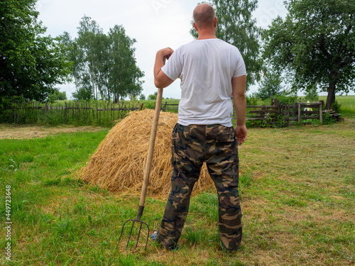 A man stands with his back to the camera with a pitchfork in front of a haystack. Rural landscape