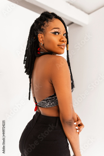 Canvas-taulu Pretty beautiful young black woman with cool dreadlocks with sexy ass in jeans in stylish bandana top with open back near white wall indoors