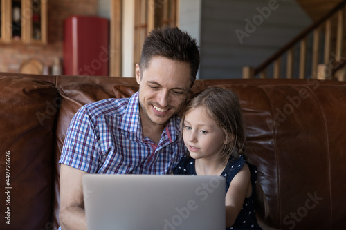 Happy father and little daughter kid using laptop at home on couch, watching online movie on computer, making video call, shopping on internet, playing virtual game, enjoying digital entertainment