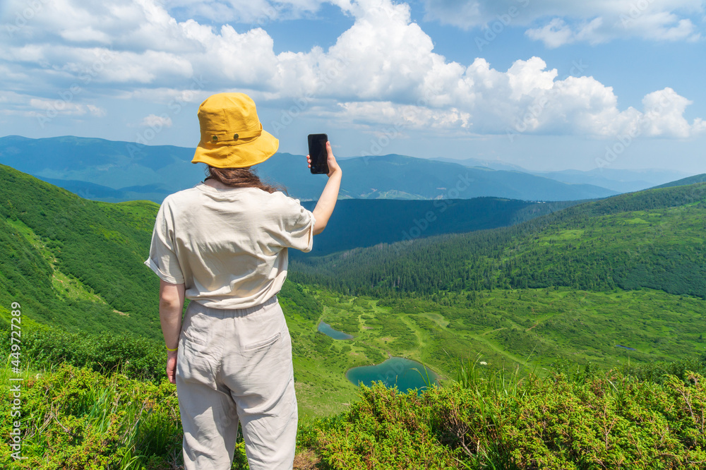 The traveler in a hat, resting in nature and using the Internet on a smartphone. A tourist girl photographs a panorama of the mountains and a mountain lake by a mobile phone. Mountain beautiful landsc