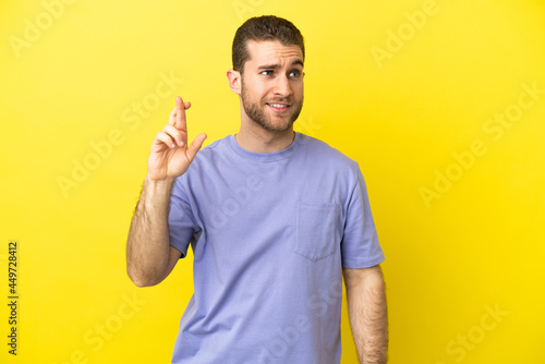 Handsome blonde man over isolated yellow background with fingers crossing and wishing the best