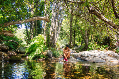 Unrecognizable Woman Sitting On A Rock Surrounded By A Spectacular Nature Landscape. Young Girl Who Is In Contact With Nature, She Is Reflected In The River.