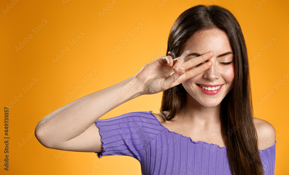 Young brunette woman smiling romantic, showing peace sign, cover eye with two fingers, standing happy against orange background Young brunette woman smiling romantic, showing peace sign, cover eye with two fingers, standing happy against orange background