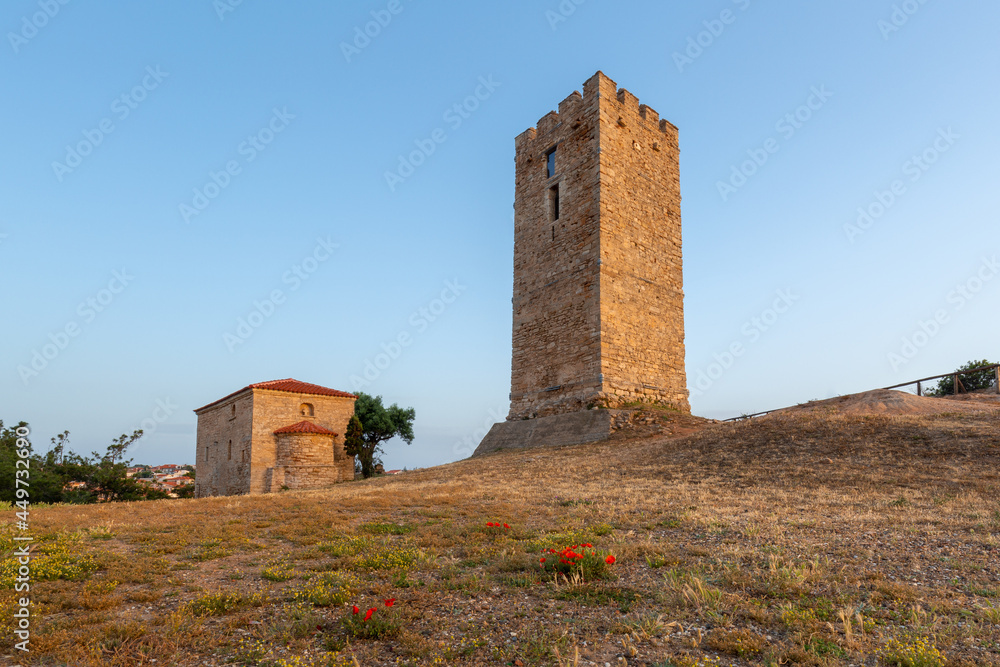 A beautifully preserved ancient Byzantine tower on a hill and a small church nearby at sunrise with bright red poppy flowers in the foreground