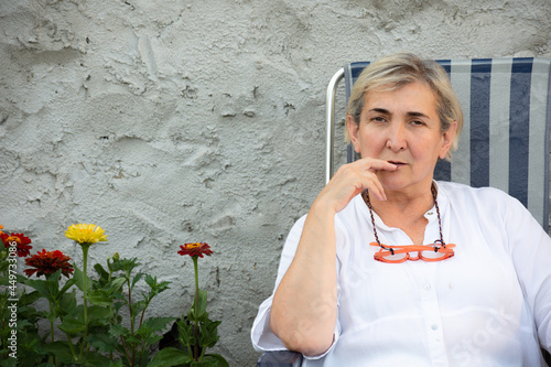 Blonde woman sit looking straight at the camera with some fingers on her mouth and her pink glasses on her neck on a summer afternoon in Spain over a grey background