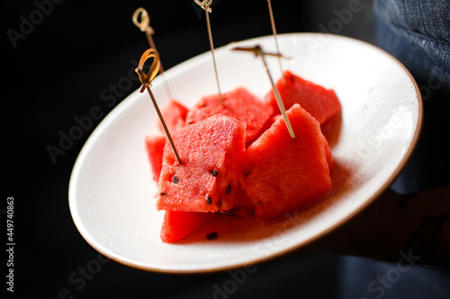 juicy watermelon sliced without peel on skewers in a white plate in the hands of a waiter on a dark background