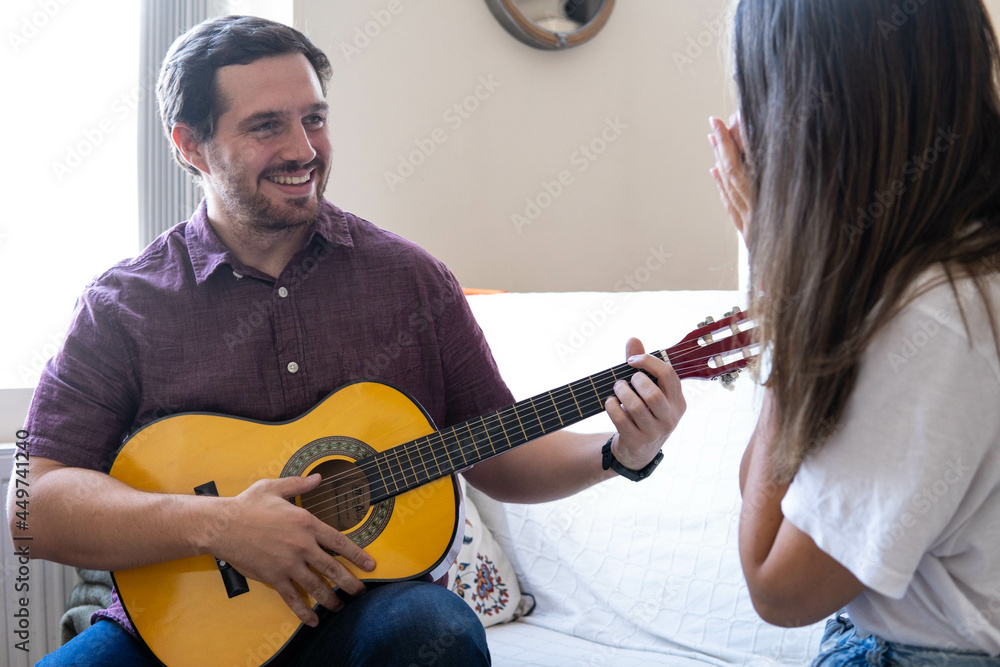 Young and attractive guy playing a romantic song in the guitar to his girlfriend while enjoying a day at home. Woman is surprised and happy listening to his song.