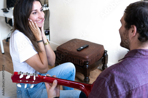 Obraz na płótnie Girl happy smiling at her boyfriend who is playing a song on the guitar for her. Romantic couple sitting on the couch at home w salonie