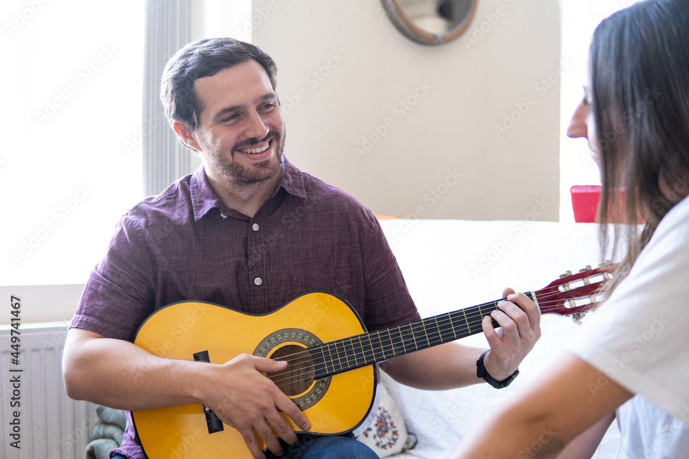 Young and attractive guy playing a romantic song in the guitar to his girlfriend while enjoying a day at home. Woman is surprised and happy listening to his song.