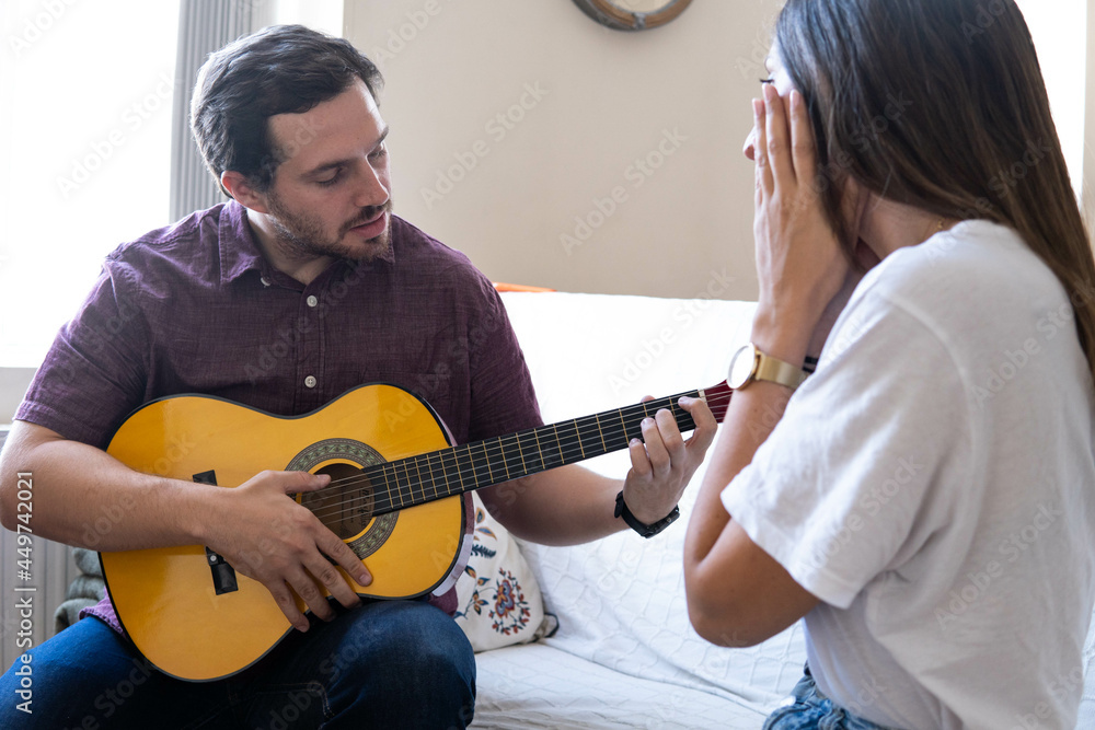 Young and attractive guy playing a romantic song in the guitar to his girlfriend while enjoying a day at home. Woman is surprised and happy listening to his song.
