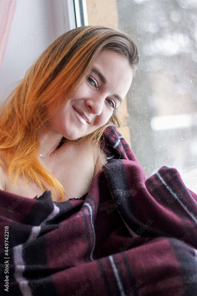a young girl with red hair sits by the window wrapped in a plaid. a young girl with red hair sits by the window wrapped in a plaid.