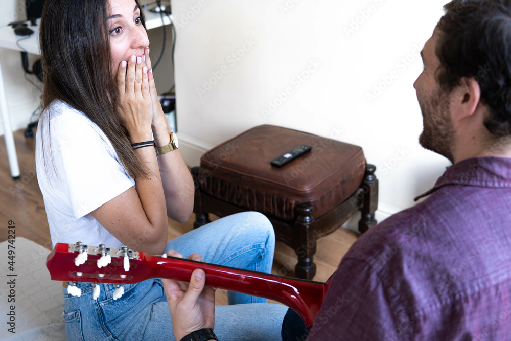 Girl happy smiling at her boyfriend who is playing a song on the guitar for her. Romantic couple sitting on the couch at home