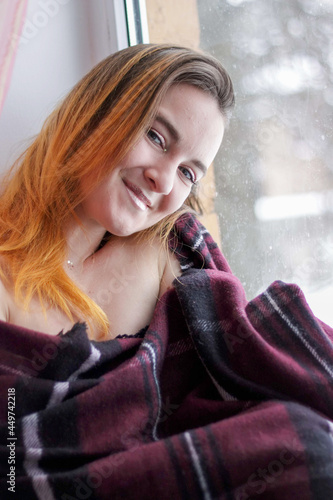 a young girl with red hair sits by the window wrapped in a plaid.