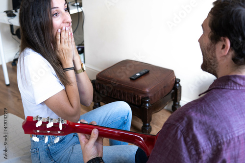Obraz na płótnie Girl happy smiling at her boyfriend who is playing a song on the guitar for her. Romantic couple sitting on the couch at home w salonie