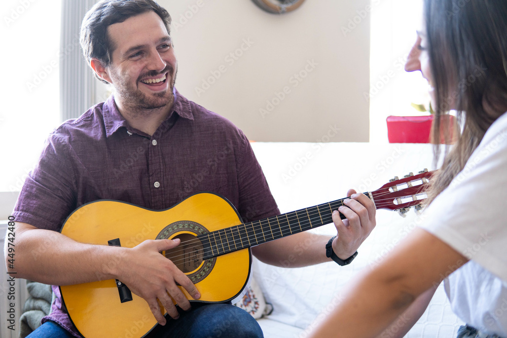 Young and attractive guy playing a romantic song in the guitar to his girlfriend while enjoying a day at home. Woman is surprised and happy listening to his song.