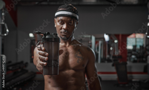 Athletic african american man with naked torso showing sports glass with water or sports nutrition on training in gym