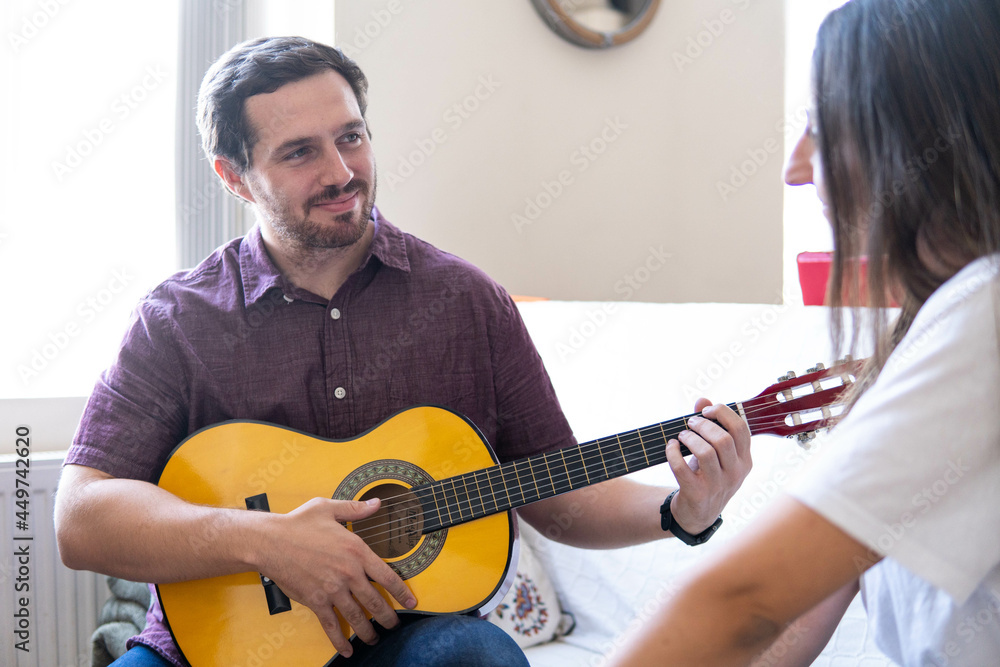 Young and attractive guy playing a romantic song in the guitar to his girlfriend while enjoying a day at home. Woman is surprised and happy listening to his song.