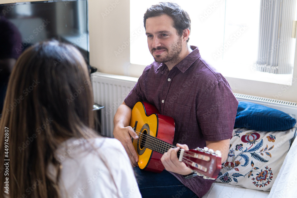 Young and attractive guy playing a romantic song in the guitar to his girlfriend while enjoying a day at home. Woman is surprised and happy listening to his song.