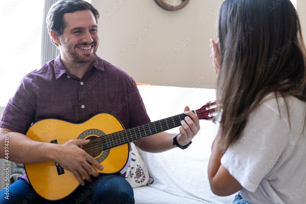 Young and attractive guy playing a romantic song in the guitar to his girlfriend while enjoying a day at home. Woman is surprised and happy listening to his song.