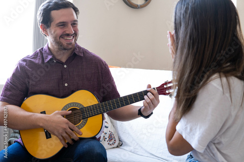 Obraz na płótnie Young and attractive guy playing a romantic song in the guitar to his girlfriend while enjoying a day at home. Woman is surprised and happy listening to his song. w salonie