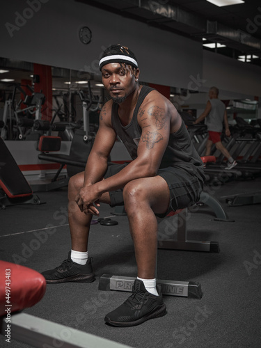 Stylish sport african american male fitness instructor sitting on bench. Handsome athletic man on training in gym
