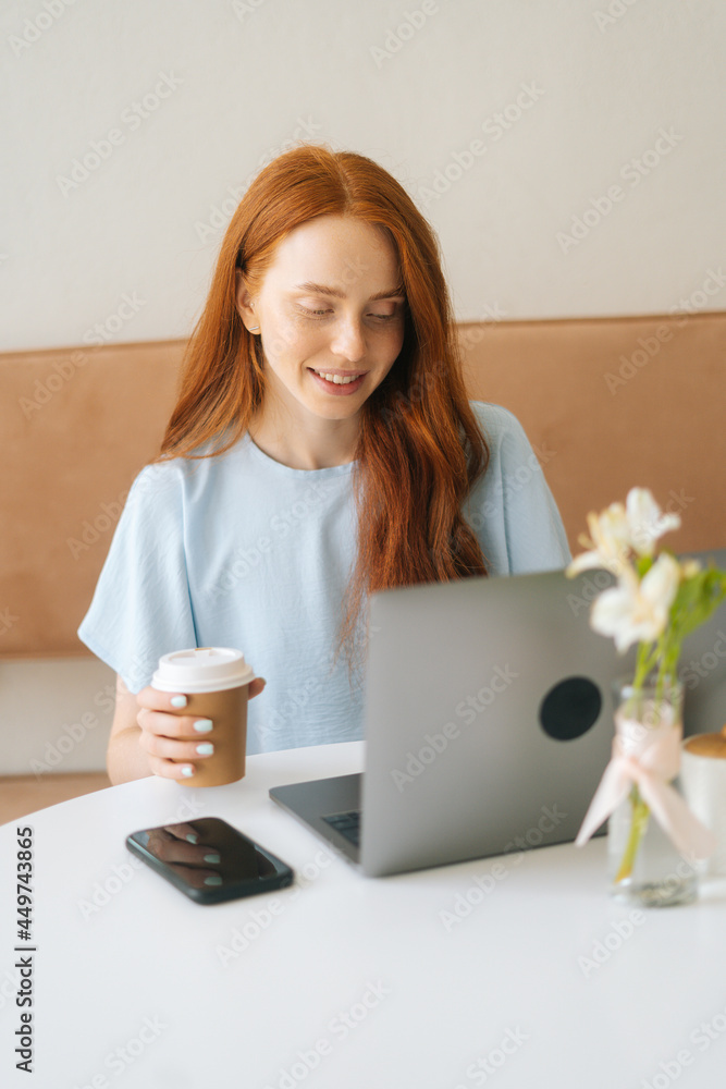 Vertical portrait of happy young beautiful woman typing on laptop and drinking coffee from cup while working remotely at cozy cafe. Pretty redhead Caucasian lady remote working or studying. Vertical portrait of happy young beautiful woman typing on laptop and drinking coffee from cup while working remotely at cozy cafe. Pretty redhead Caucasian lady remote working or studying.