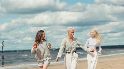 Slow motion shot of two joyful young women walking along sea coast on beach. Happy girlfriend running to join them and taking piggyback ride. Females feeling carefree and happy celebrating life