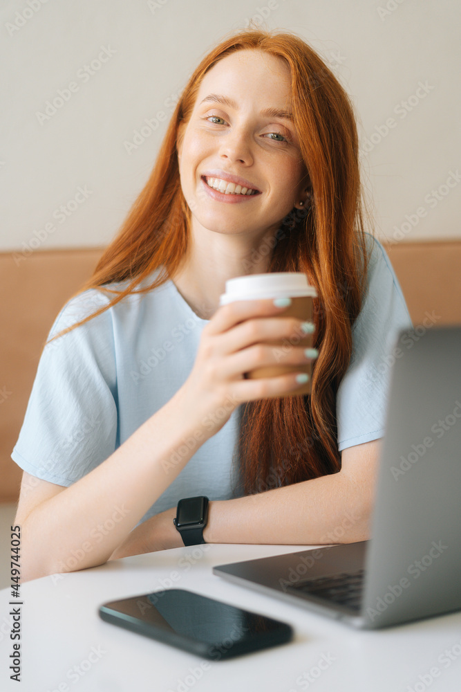 Close-up vertical portrait of happy young beautiful woman holding cup with hot coffee sitting at desk with laptop at cafe, looking at camera. Pretty redhead Caucasian lady remote working or studying. Close-up vertical portrait of happy young beautiful woman holding cup with hot coffee sitting at desk with laptop at cafe, looking at camera. Pretty redhead Caucasian lady remote working or studying.